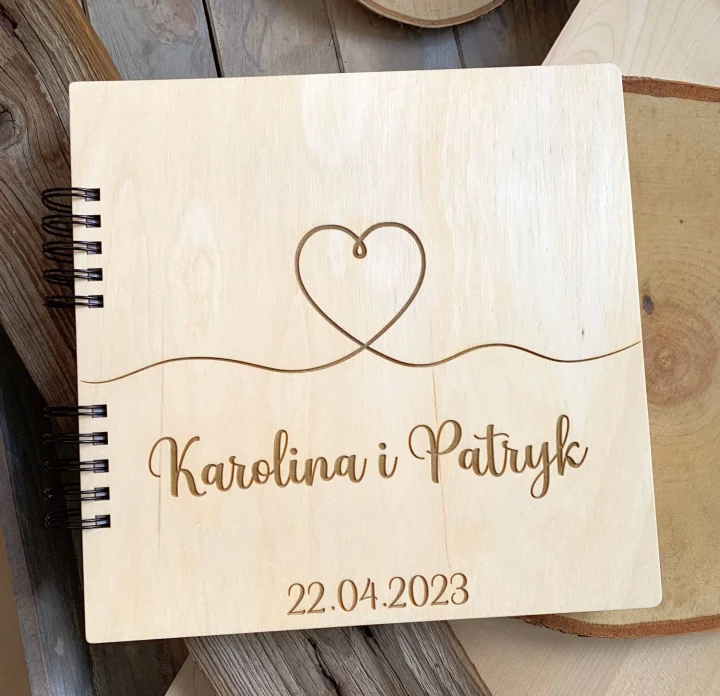 Drewniany album na zdjęcia z grawerem serca "one line", imionami Karolina i Patryk oraz datą, leżący na plastrach drewna.