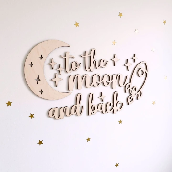 Zestaw dekoracji ściennych wyciętych z jasnej, naturalnej sklejki. W skład zestawu wchodzi duży ażurowy księżyc, napis 'to the moon and back' wykonany ozdobną czcionką, rakieta oraz komplet małych gwiazdek. Produkt prezentowany na neutralnym, jasnym tle.