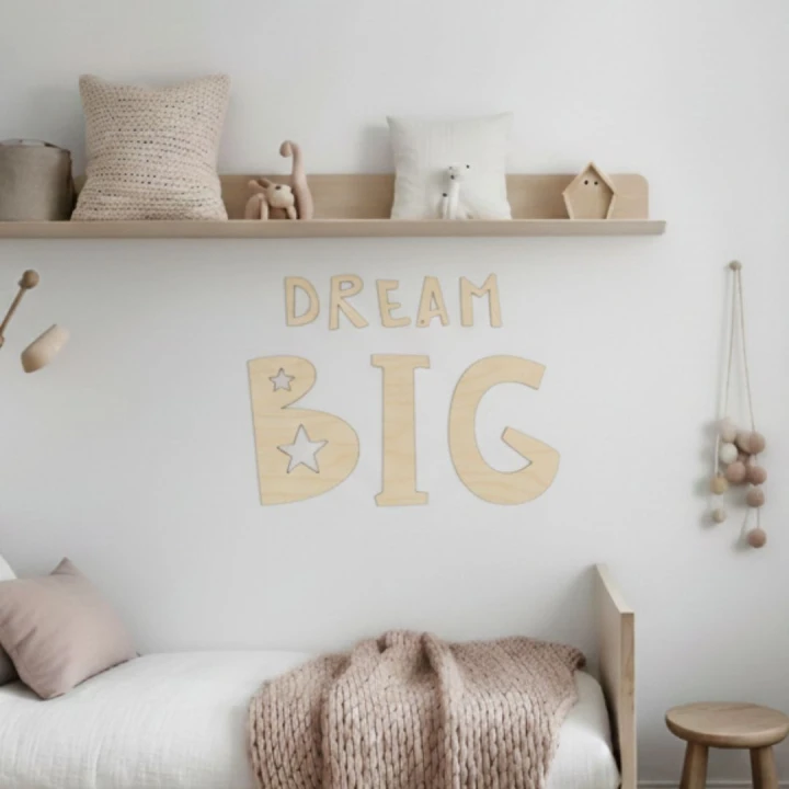 Duży napis ze sklejki DREAM BIG zamontowany nad łóżeczkiem w jasnym pokoju dziecięcym, obok wiszące drewniane gwiazdki, styl minimalistyczny.