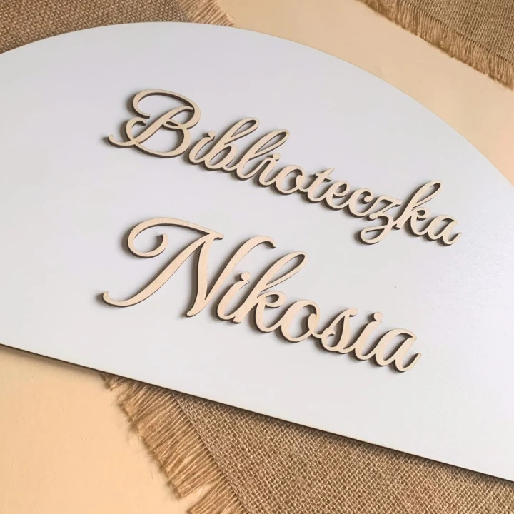Biała tablica HDF w kształcie półkola z naklejonym drewnianym napisem Biblioteczka z imieniem.