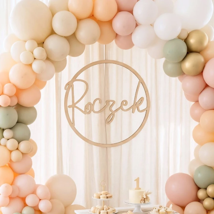 Dekoracja Roczek ze sklejki zawieszona na ściance balonowej w jasnych, pastelowych kolorach.