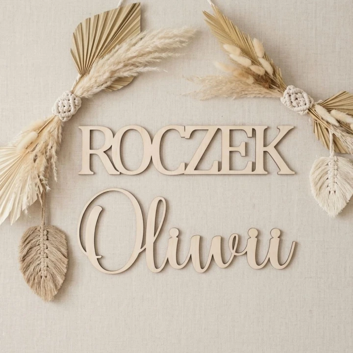 Drewniany napis dekoracyjny na roczek z imieniem Oliwia ułożony na lnianym tle w otoczeniu suchych traw i makramy.