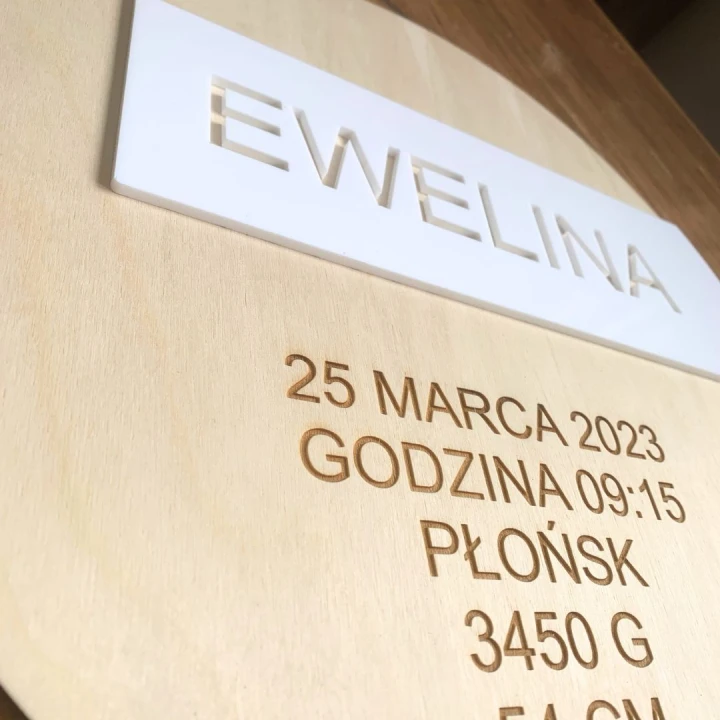 Okrągła drewniana metryczka urodzenia dla dziecka . Zbliżenie na imię wycięte w prostokątnej, białej plexi (efekt 3D)