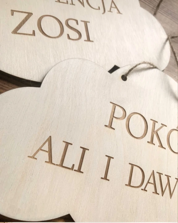 Detal personalizowanej drewnianej tabliczki ze sklejki w kształcie chmurki z wygrawerowanym napisem na drzwi dziecięce. Zbliżenie ukazuje naturalny słój drewna oraz ciemniejszy, czysty grawer.